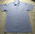 A light blue short-sleeve button-up shirt with a subtle grid pattern, laid flat on a beige shag rug. The shirt features a classic collar, a chest pocket, and a button-down front.