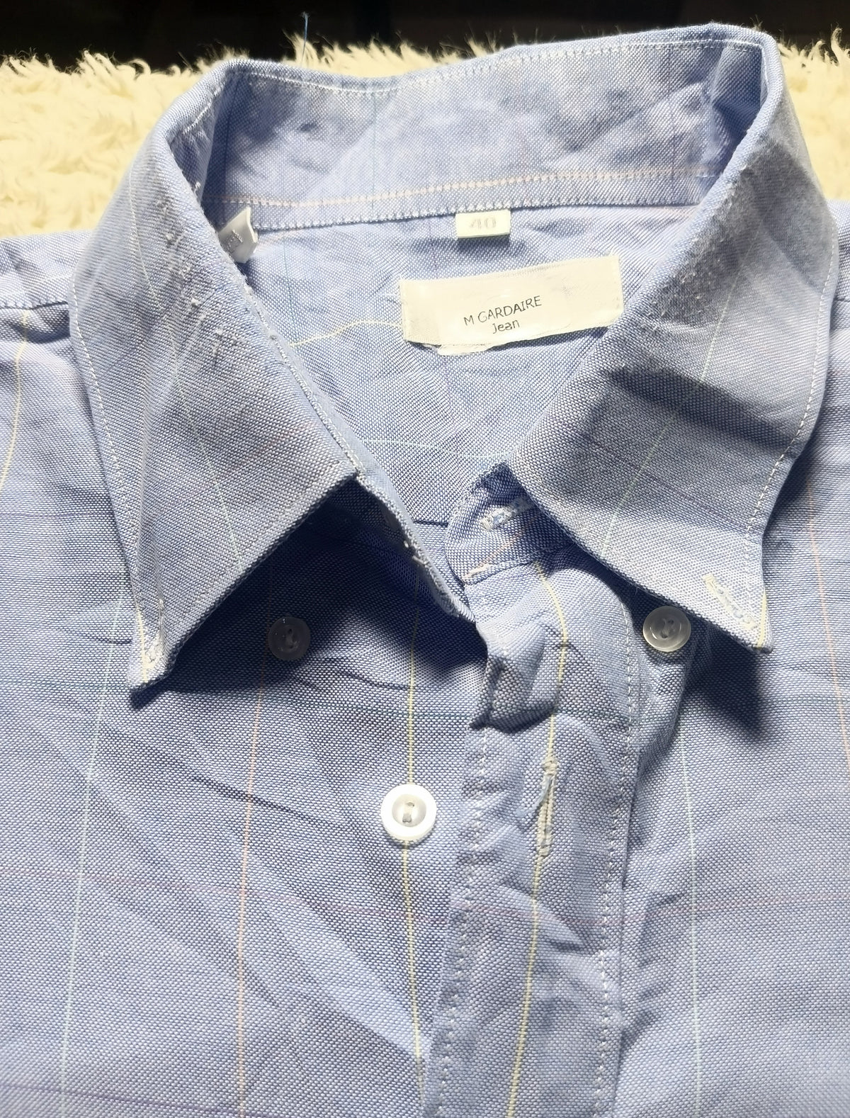 A close-up of a light blue button-down shirt with thin vertical pinstripes, laid flat on a textured beige surface. The brand label inside the collar reads 'MY CLOSET'.