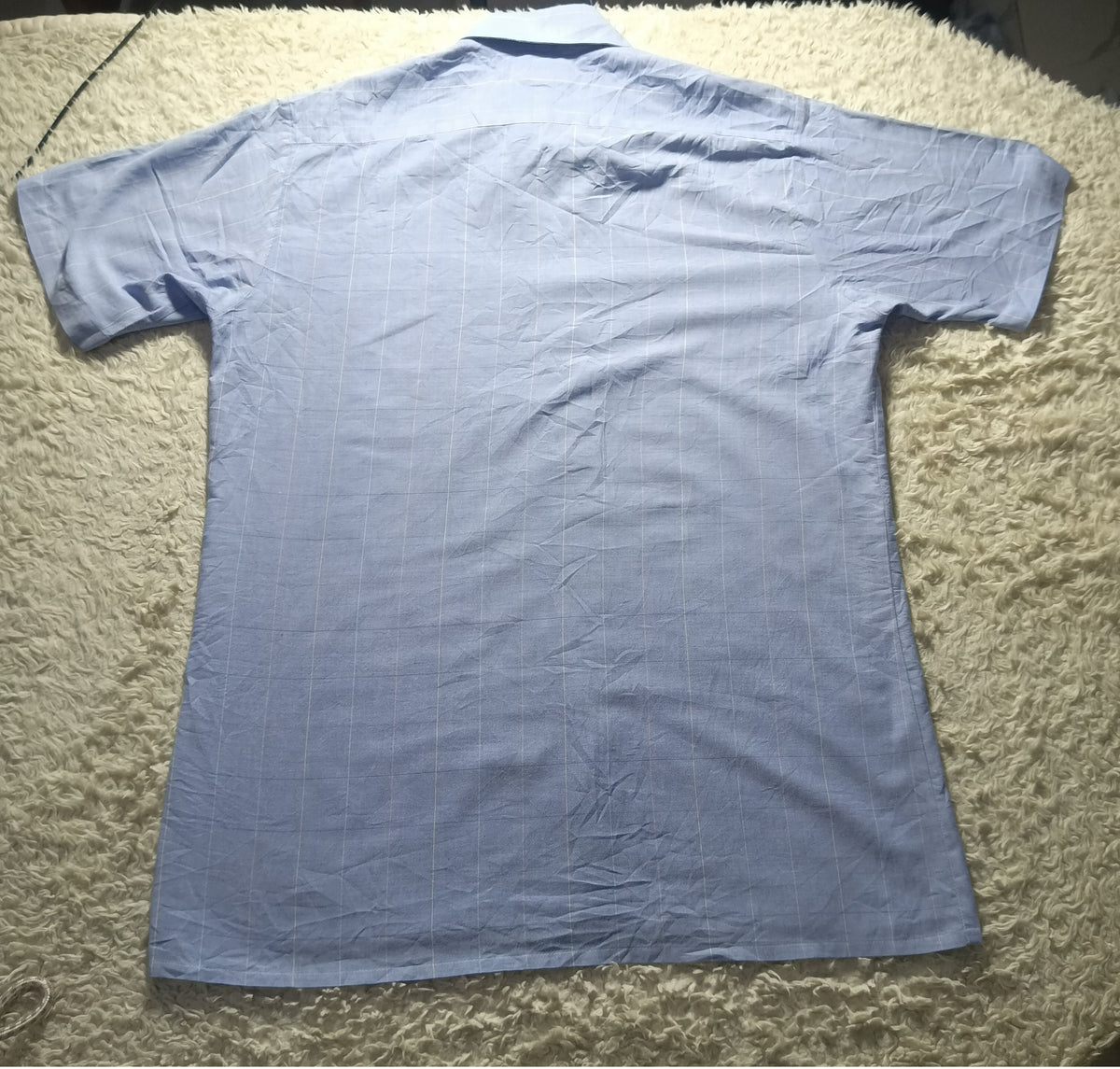 A light blue short-sleeve button-up shirt laid flat on a beige shag rug, viewed from the back. The photo has camera metadata text overlaid at the bottom.