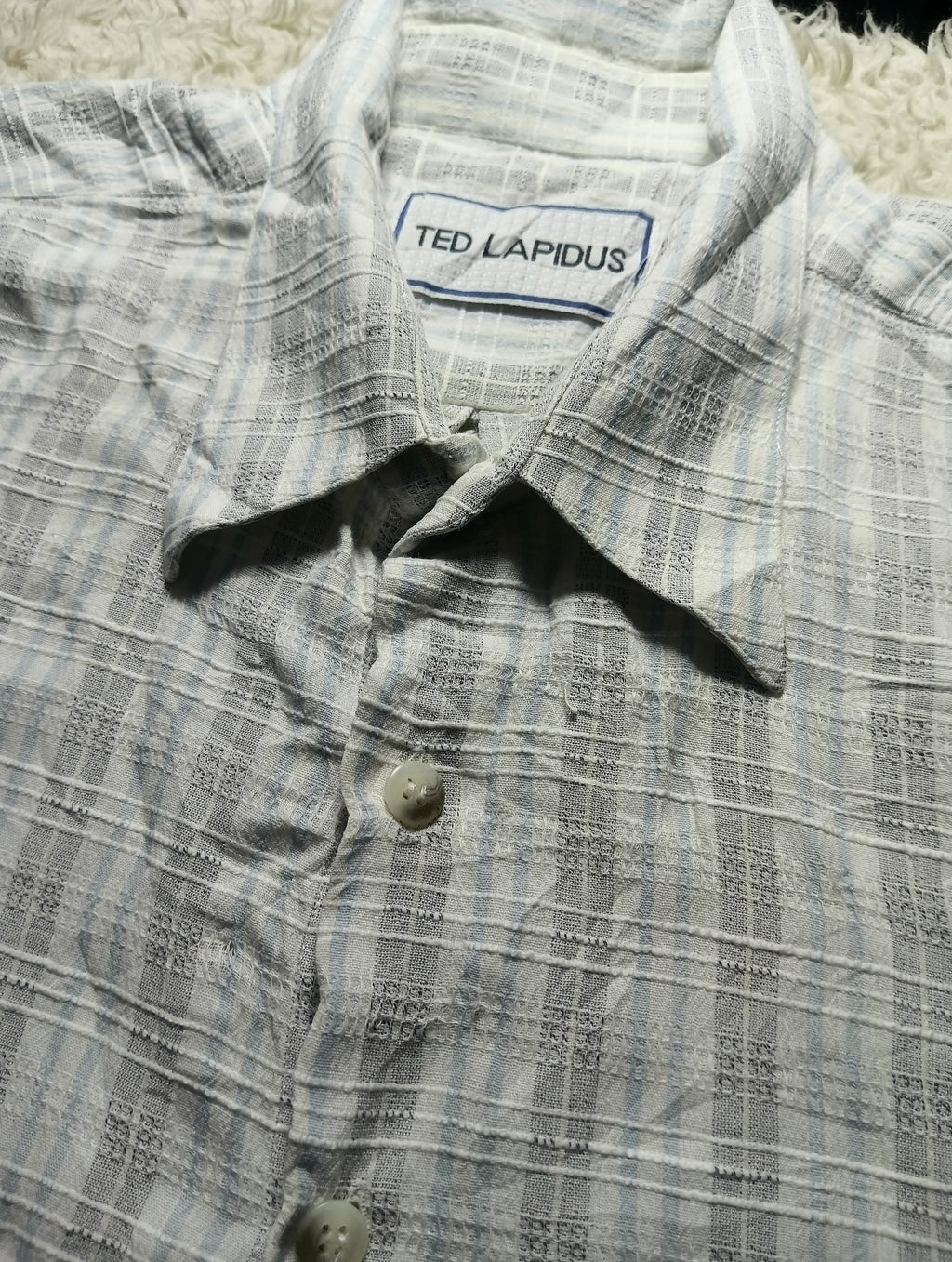A close-up of a Ted Lapidus button-up shirt with a textured, woven plaid pattern in light gray and white. The brand label 'TED LAPIDUS' is visible inside the collar.