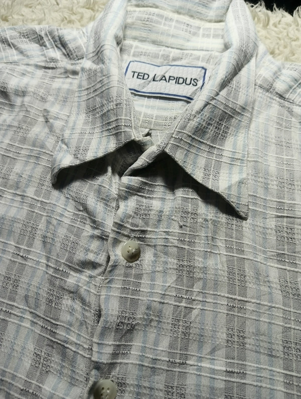 A close-up of a Ted Lapidus button-up shirt with a textured, woven plaid pattern in light gray and white. The brand label 'TED LAPIDUS' is visible inside the collar.