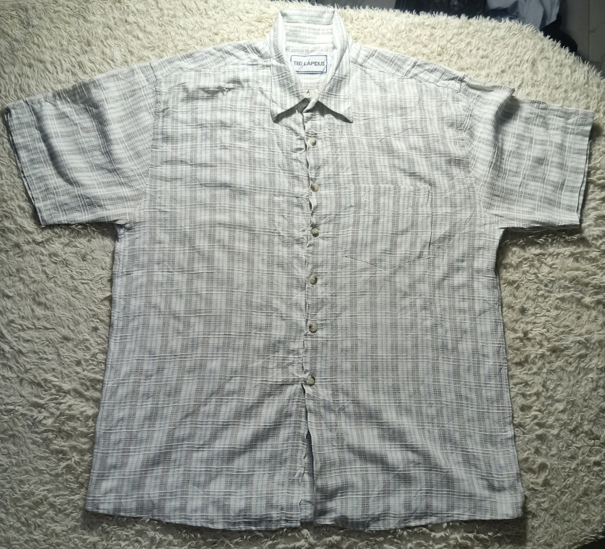 A short-sleeve button-up shirt with a fine vertical stripe pattern in gray and white, laid flat on a textured beige shag rug. The brand name 'SOLIS' is visible on the inner neck label.