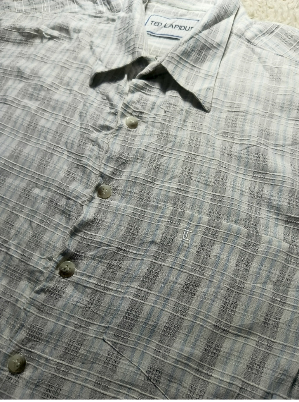 A close-up of a Ted Lapidus short-sleeve button-up shirt in a gray and white plaid pattern, laid flat on a light beige surface.