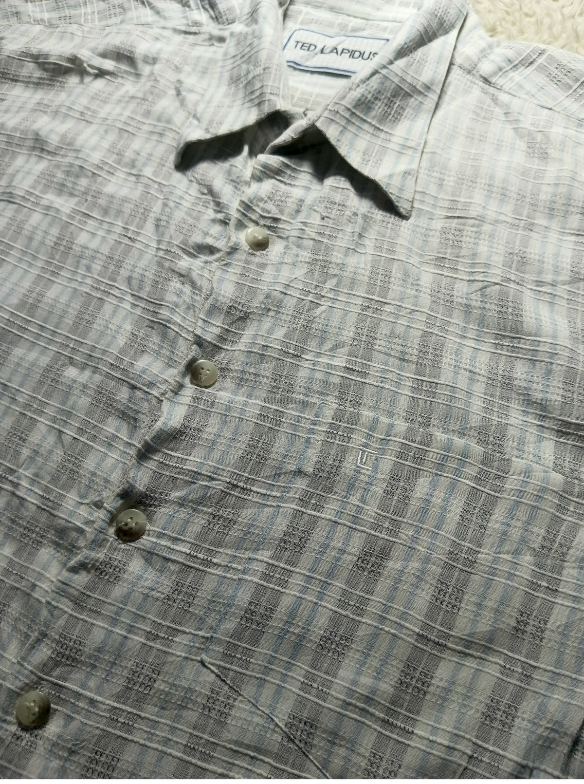 A close-up of a Ted Lapidus short-sleeve button-up shirt in a gray and white plaid pattern, laid flat on a light beige surface.