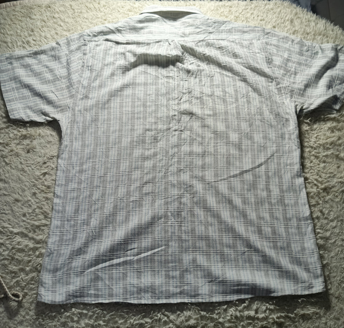 A short-sleeve button-up shirt with a gray and white plaid pattern, laid flat on a textured gray shag rug.