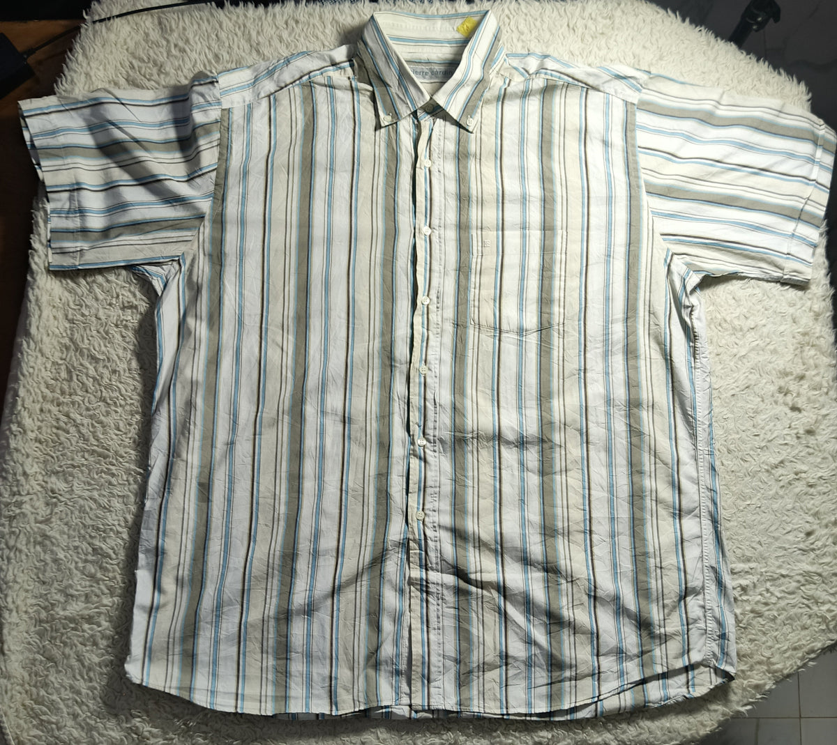 A short-sleeve button-up shirt with vertical stripes in white, blue, and green, laid flat on a white textured blanket.