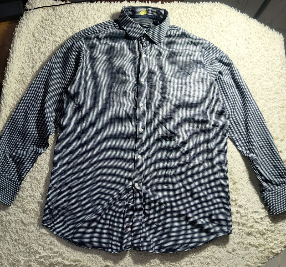 A long-sleeve button-up shirt in a faded blue-gray color, laid flat on a white shaggy blanket in a home setting.