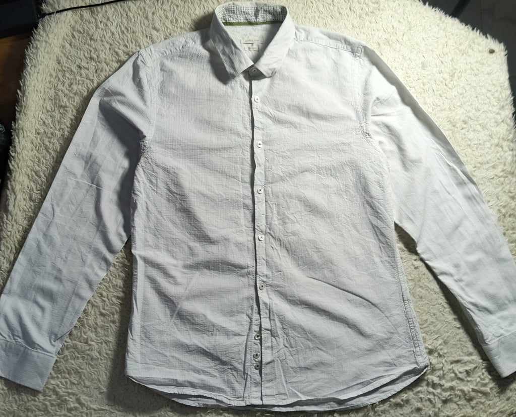 A white long-sleeve button-up shirt laid flat on a white shaggy blanket, photographed in a casual indoor setting with some clutter visible around it.