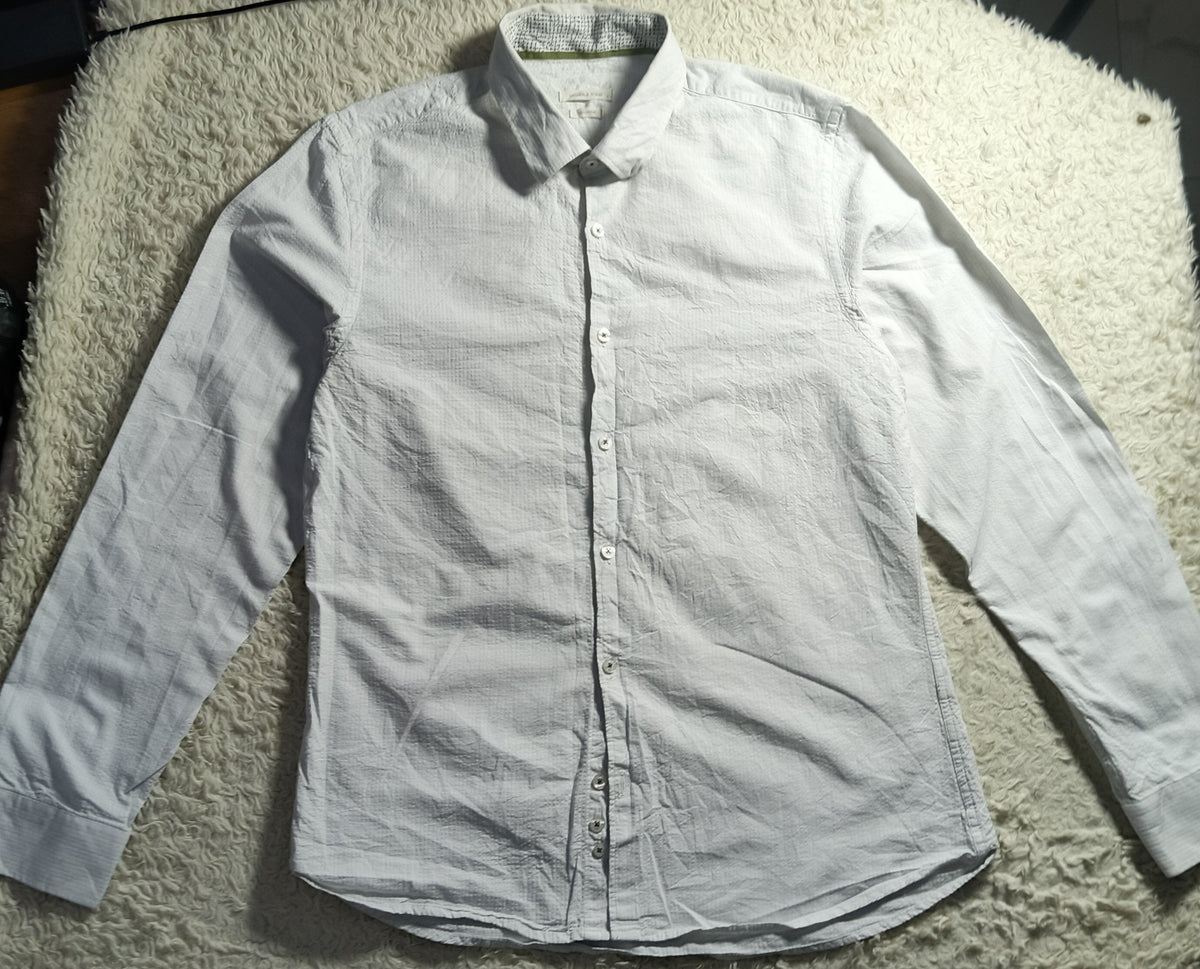 A white long-sleeve button-up shirt laid flat on a white shaggy blanket, photographed in a casual indoor setting with some clutter visible around it.