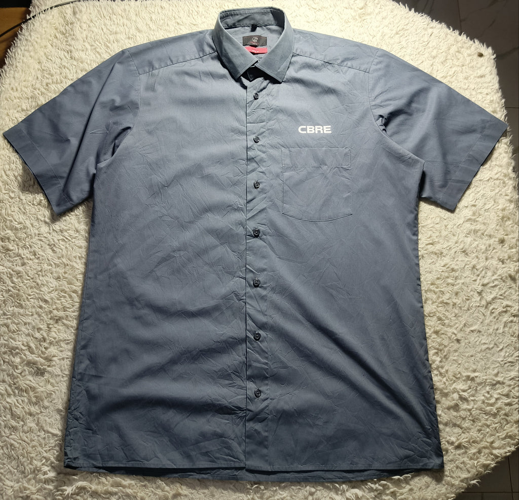 A short-sleeve button-up shirt in a muted slate blue-gray color, laid flat on a white shag rug. The shirt features a small white 'CARE' logo on the left chest.