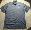 A short-sleeve button-up shirt in a muted slate blue-gray color, laid flat on a white shag rug. The shirt features a small white 'CARE' logo on the left chest.