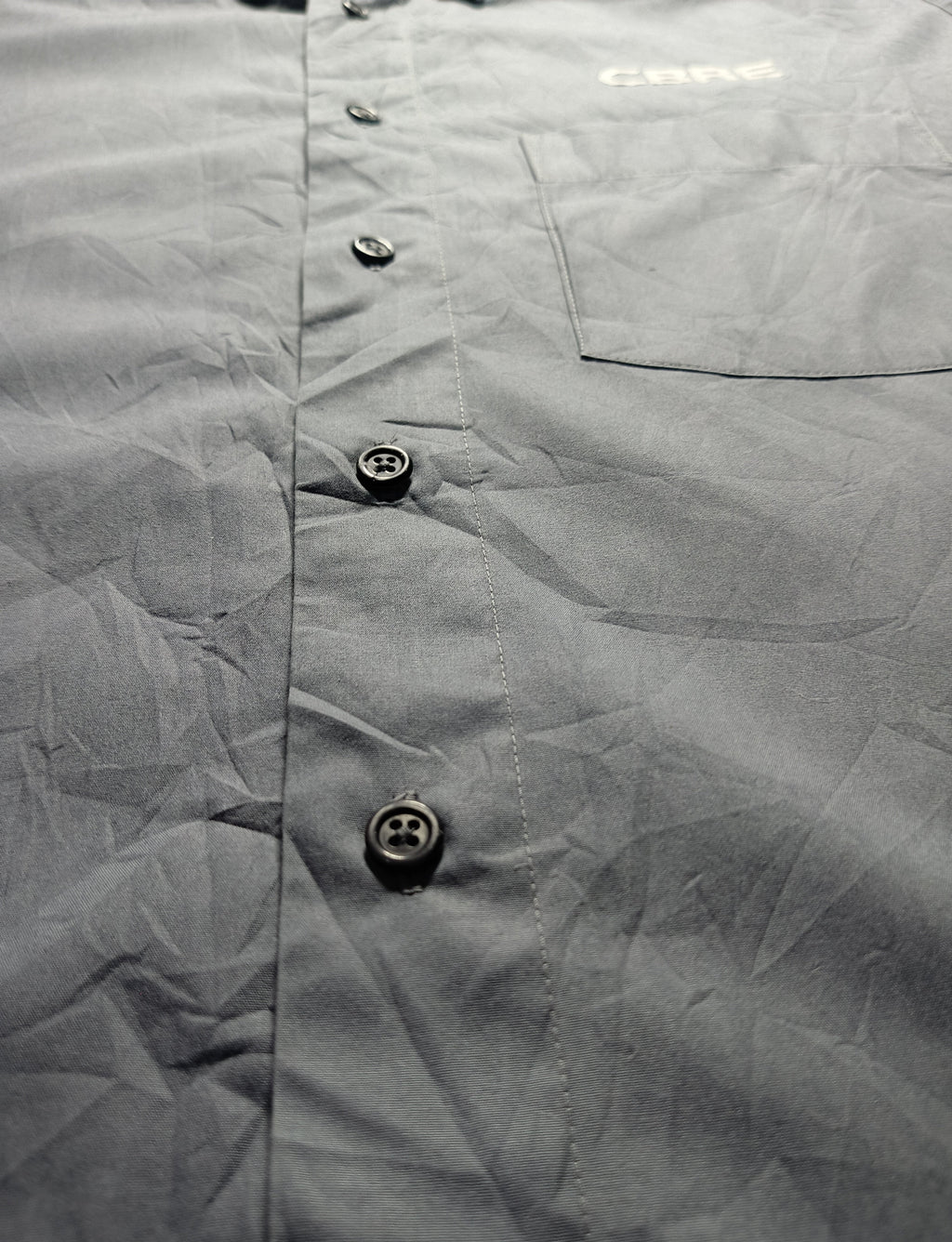 Close-up of a gray button-up shirt fabric with a chest pocket and visible 'CAMEO' branding near the collar area, photographed against a dark background.