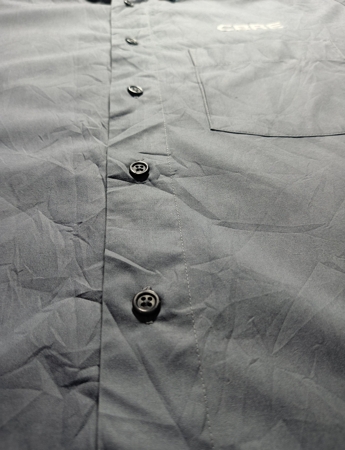 Close-up of a gray button-up shirt fabric with a chest pocket and visible 'CAMEO' branding near the collar area, photographed against a dark background.