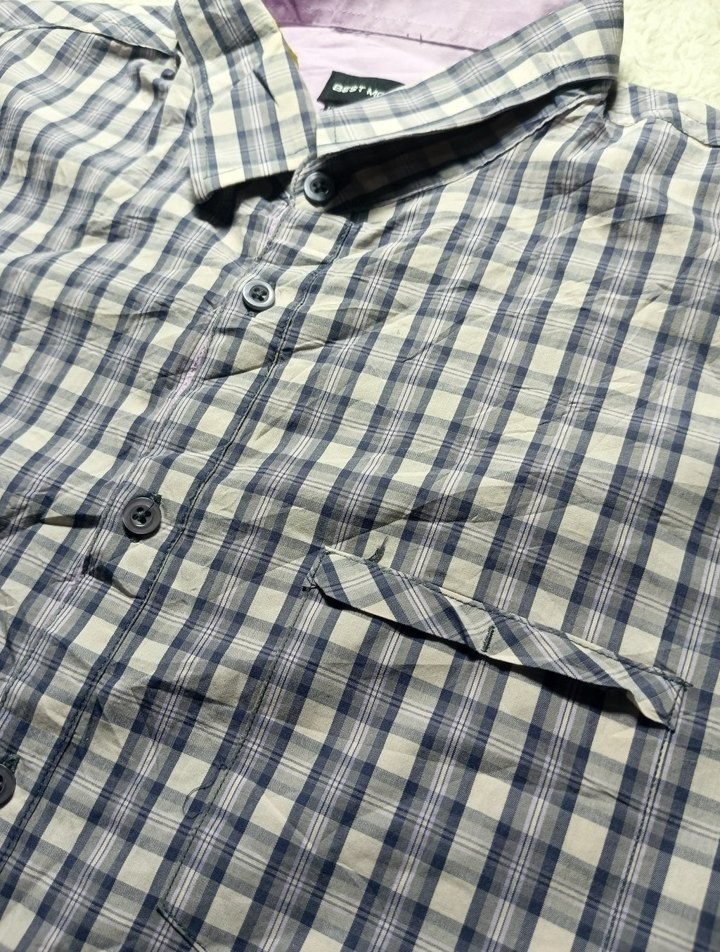 A close-up of a plaid button-up shirt with a blue, gray, and beige checkered pattern, featuring a collar, dark buttons, and a chest pocket.