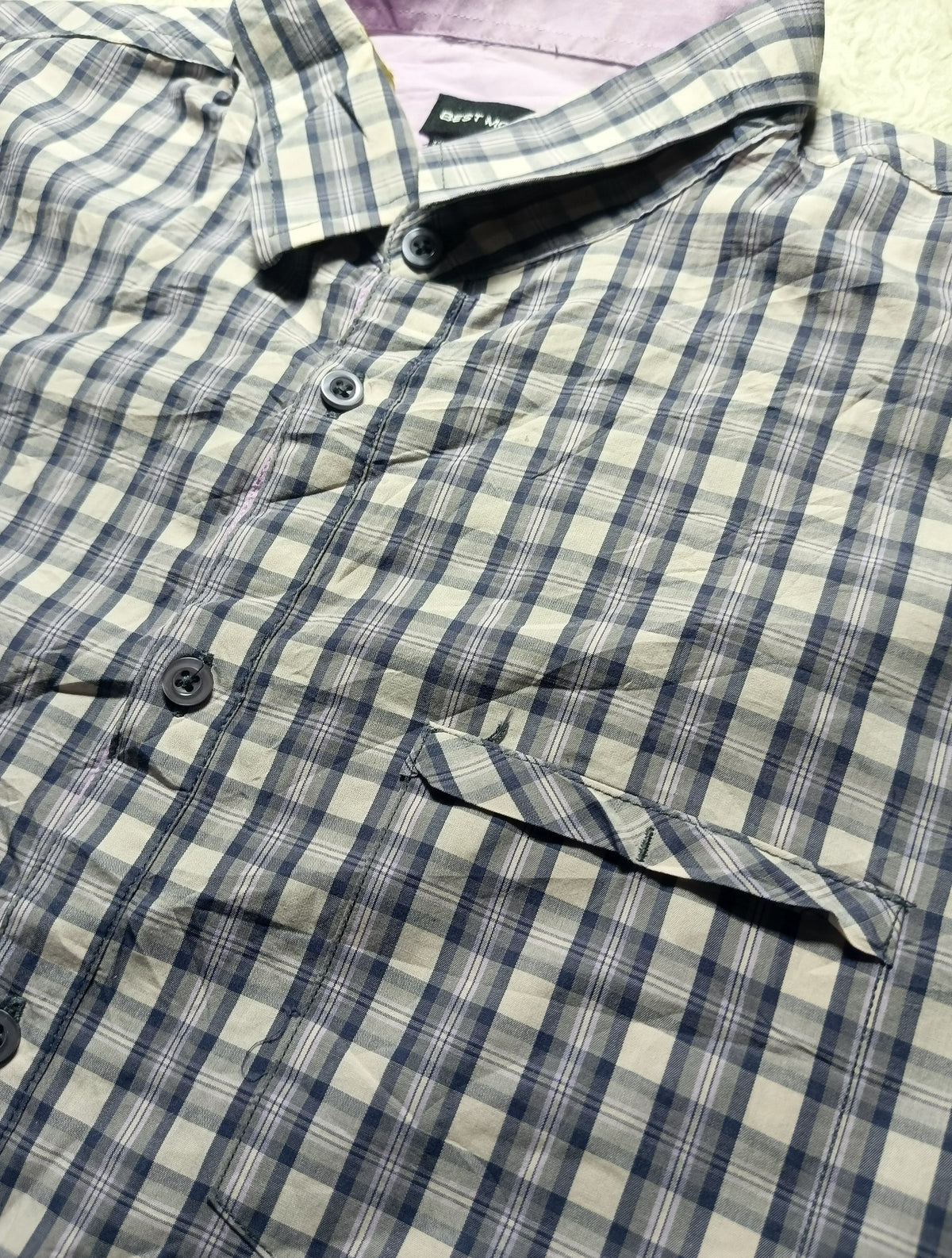 A close-up of a plaid button-up shirt with a blue, gray, and beige checkered pattern, featuring a collar, dark buttons, and a chest pocket.