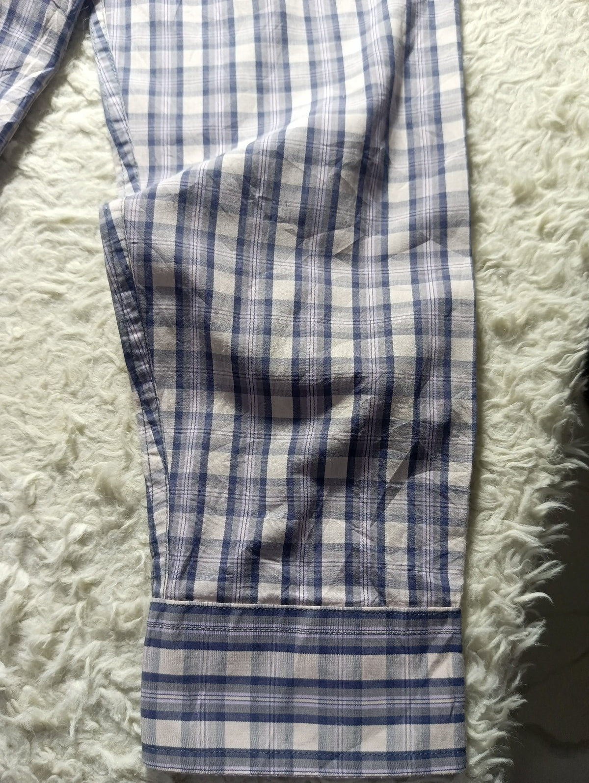 Blue and white checkered pajama pants laid flat on a white shaggy blanket, with the cuff showing a buttoned pocket.