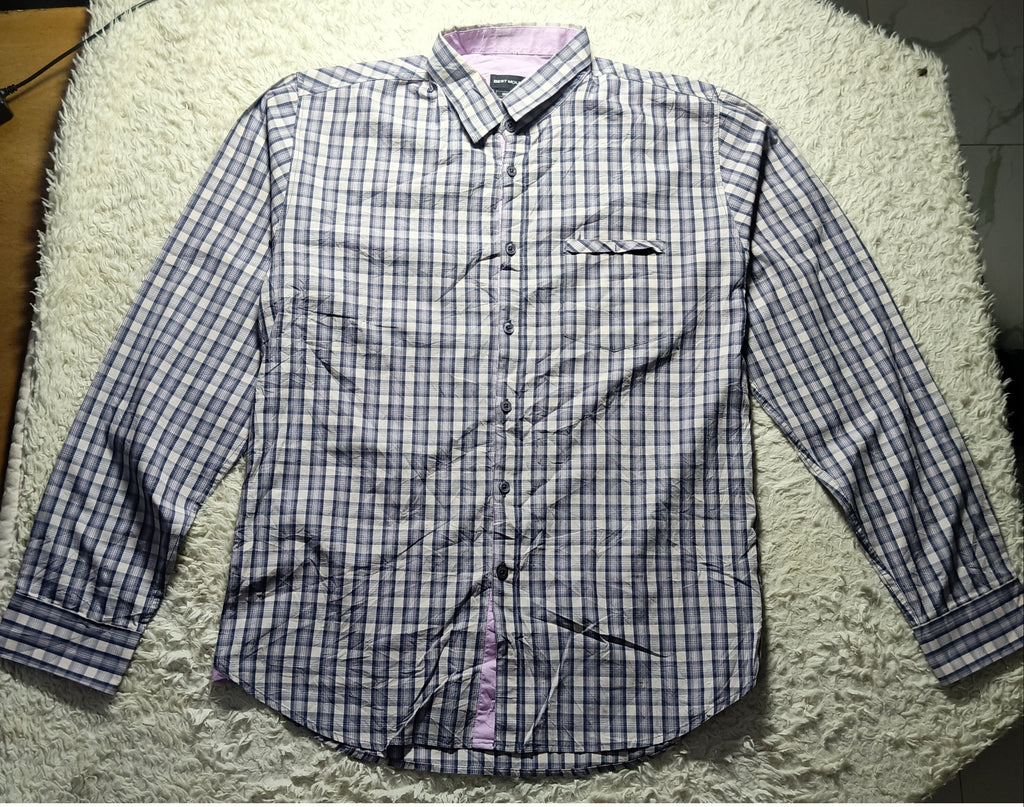 A long-sleeve button-up shirt with a blue and white checkered pattern, laid flat on a white shaggy rug. The shirt features a single chest pocket and a light purple inner collar lining.