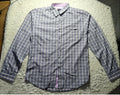 A long-sleeve button-up shirt with a blue and white checkered pattern, laid flat on a white shaggy rug. The shirt features a single chest pocket and a light purple inner collar lining.