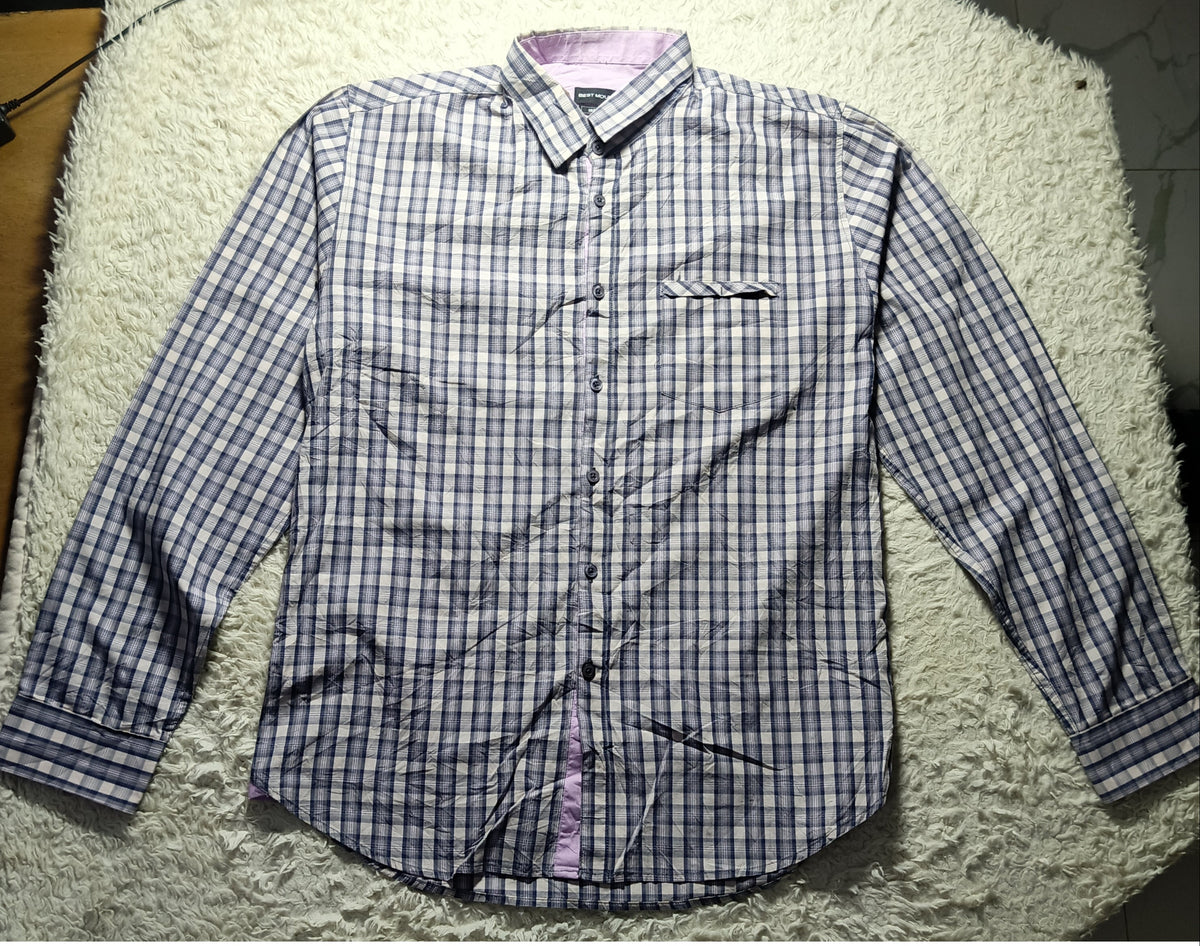A long-sleeve button-up shirt with a blue and white checkered pattern, laid flat on a white shaggy rug. The shirt features a single chest pocket and a light purple inner collar lining.