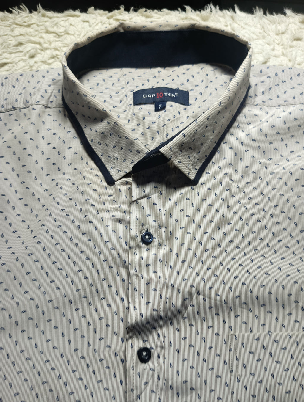 A close-up of a men's button-down shirt by Capstone, featuring a light beige base with a small blue leaf pattern and contrasting dark navy trim on the collar and front buttons.