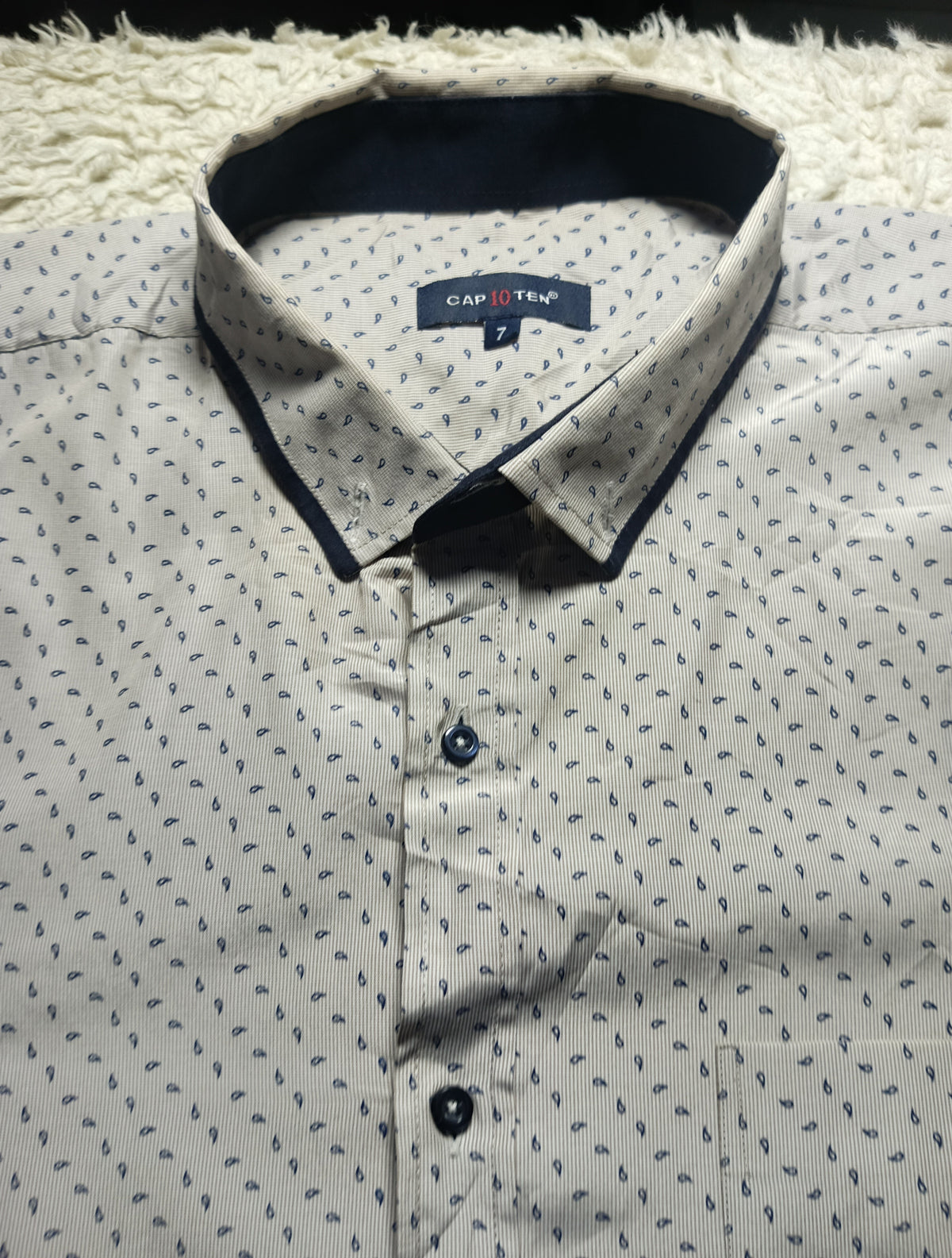 A close-up of a men's button-down shirt by Capstone, featuring a light beige base with a small blue leaf pattern and contrasting dark navy trim on the collar and front buttons.