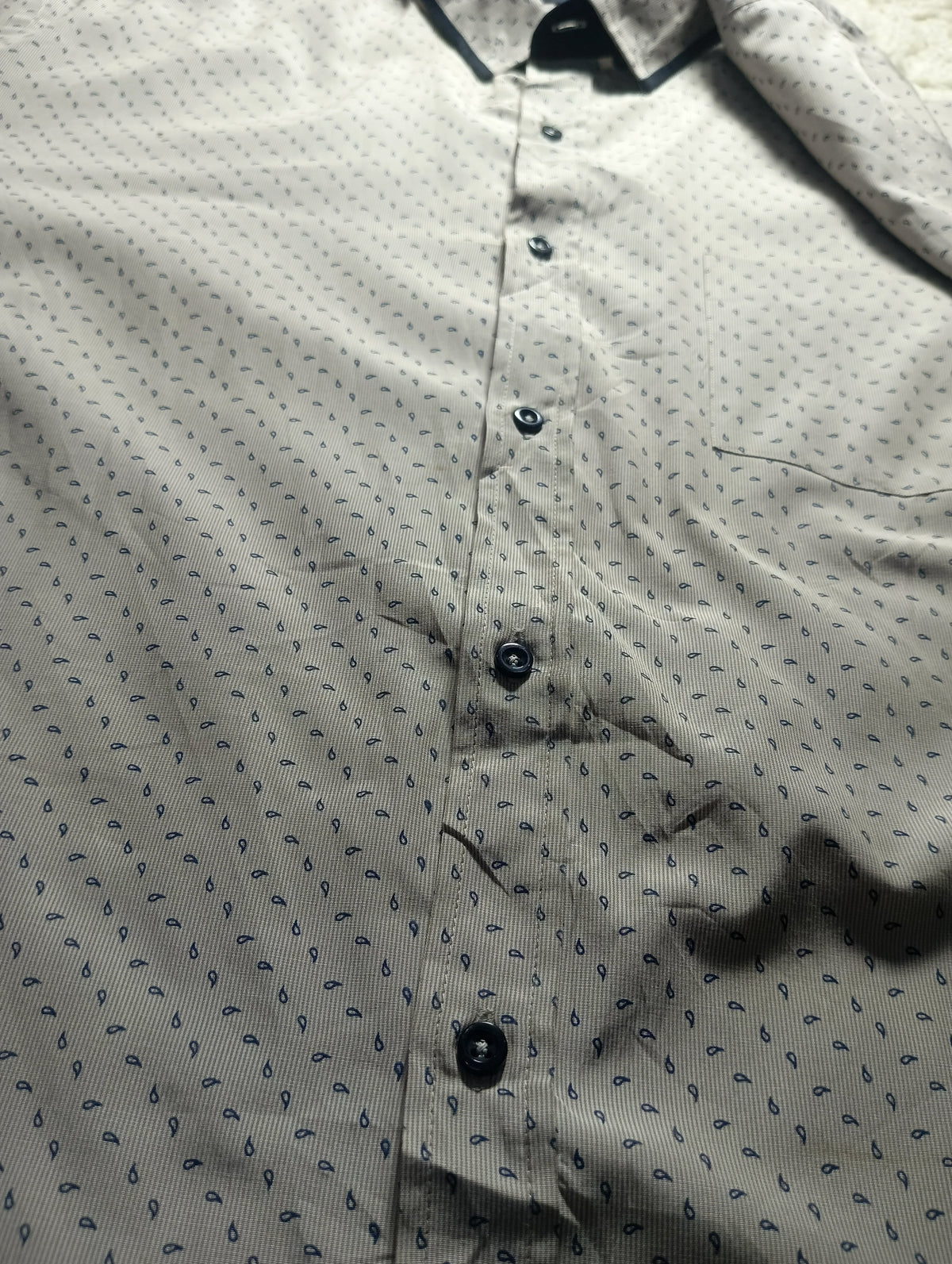 A close-up of a light beige or off-white button-up shirt with a small repeating dark blue leaf or teardrop pattern, featuring dark buttons and a dark collar.