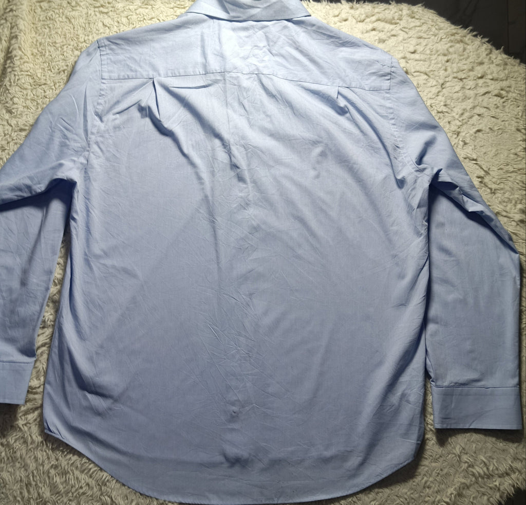 A light blue long-sleeve button-up shirt laid flat on a white shaggy blanket, photographed in a home setting with a chair and floor visible in the background.
