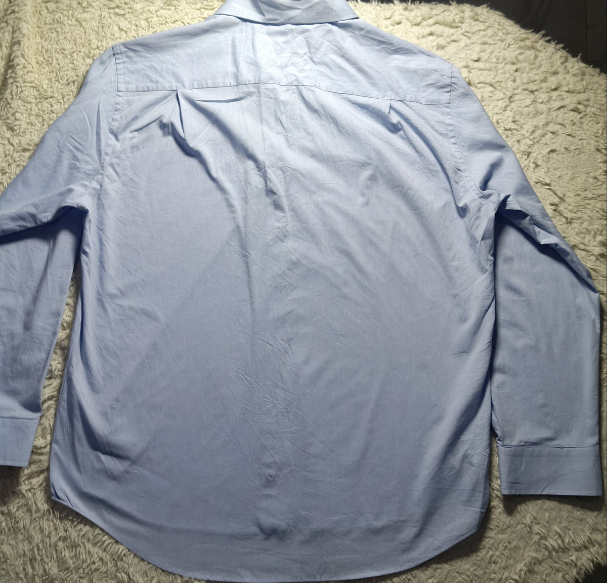 A light blue long-sleeve button-up shirt laid flat on a white shaggy blanket, photographed in a home setting with a chair and floor visible in the background.