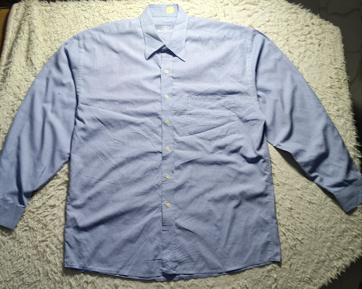 A light blue long-sleeve button-up shirt laid flat on a white shaggy blanket, photographed indoors on a tiled floor.