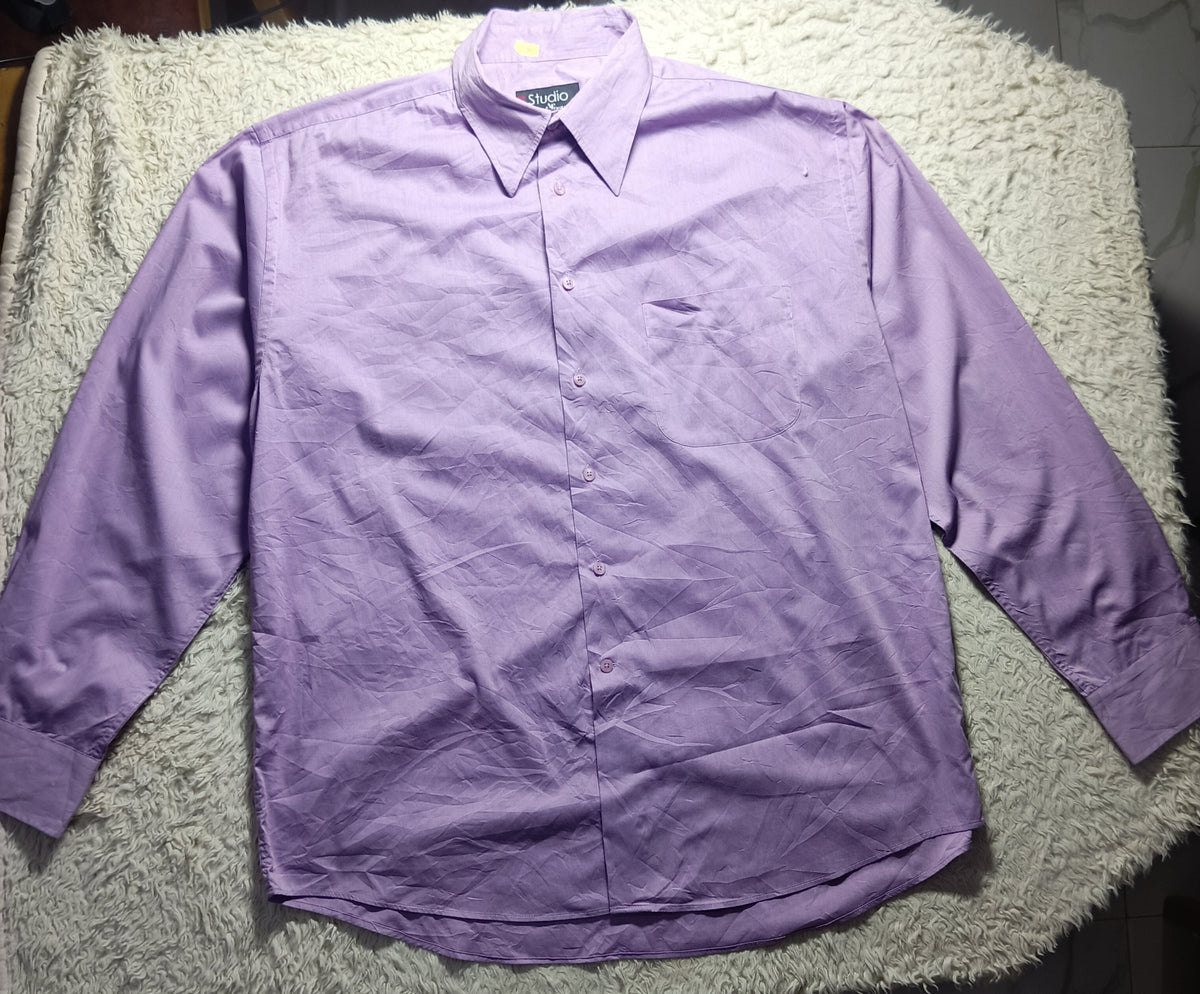 A light purple long-sleeve button-up shirt laid flat on a white shaggy blanket, with a tiled floor and dark furniture visible in the background.