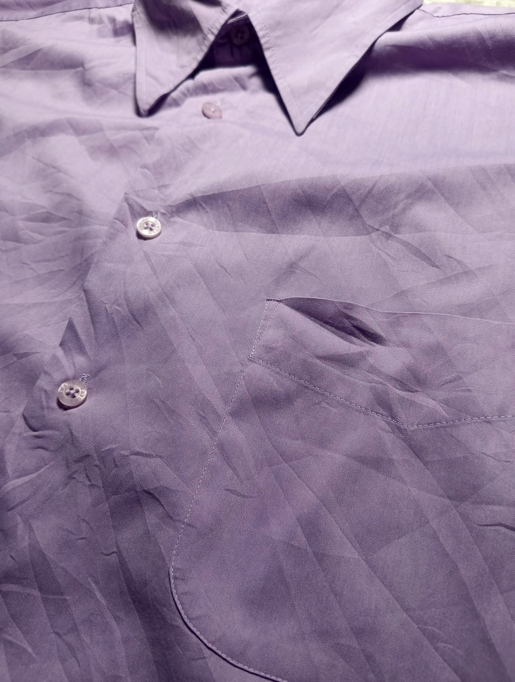 A close-up of a wrinkled lavender-purple button-down shirt, showing the collar, front buttons, and chest pocket.