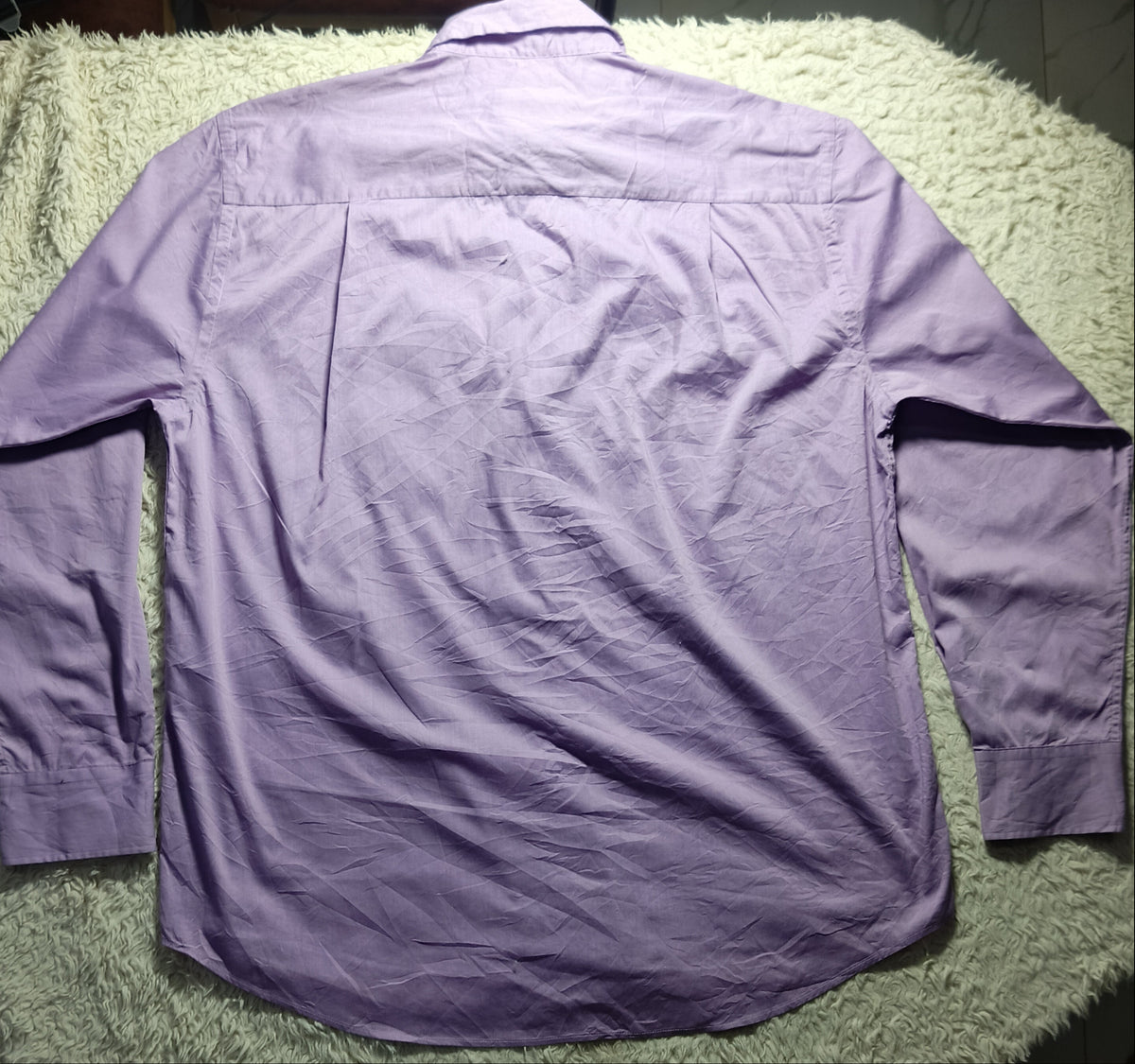 A lavender purple long-sleeve button-up shirt laid flat on a white shaggy rug, photographed from above in a home setting.
