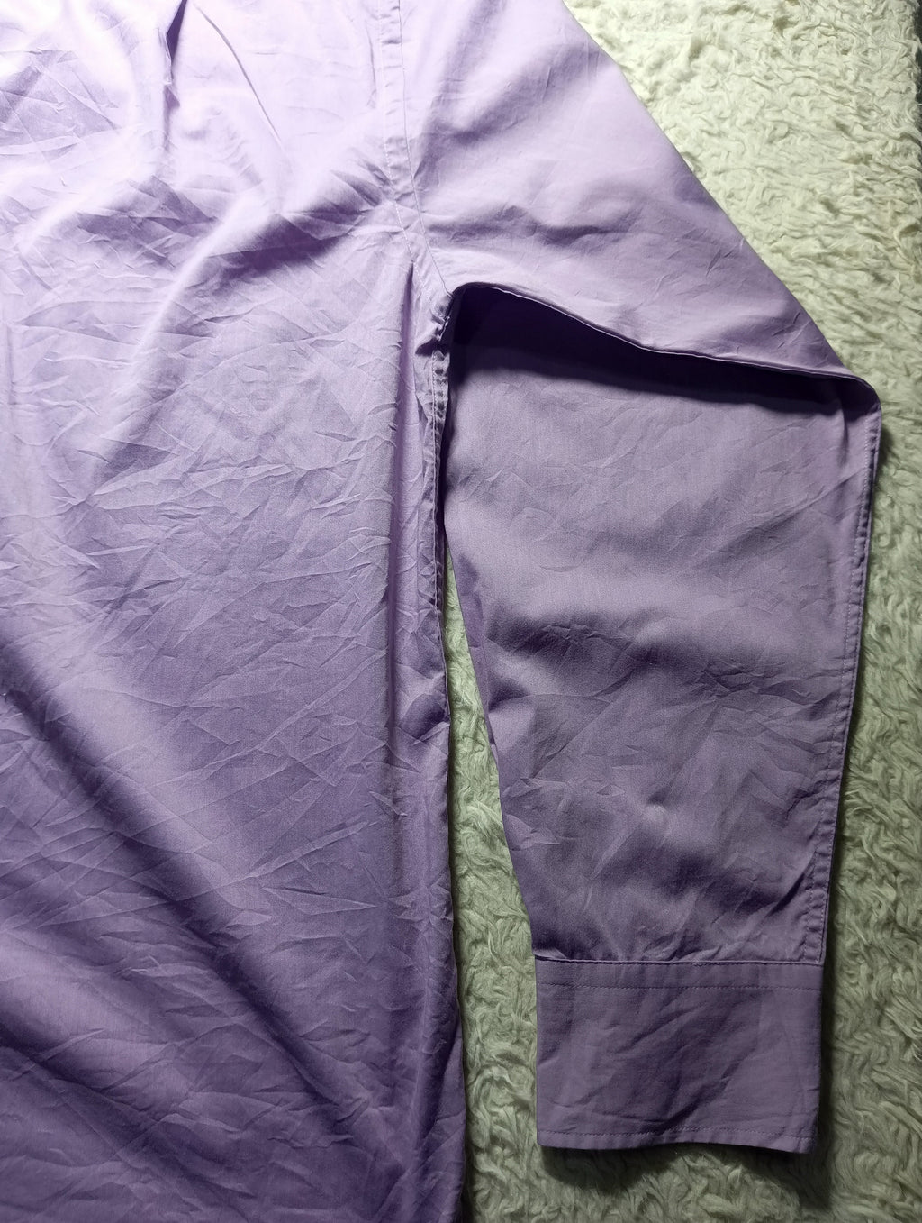 A lavender purple long-sleeve shirt laid flat on a textured green surface, showing the sleeve and body fabric.