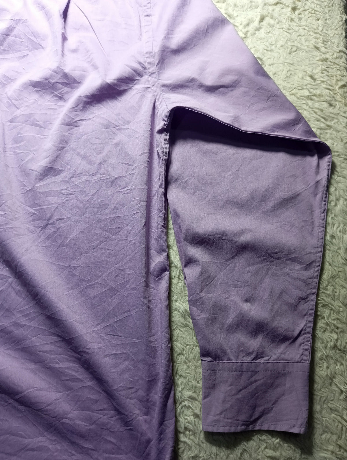 A lavender purple long-sleeve shirt laid flat on a textured green surface, showing the sleeve and body fabric.