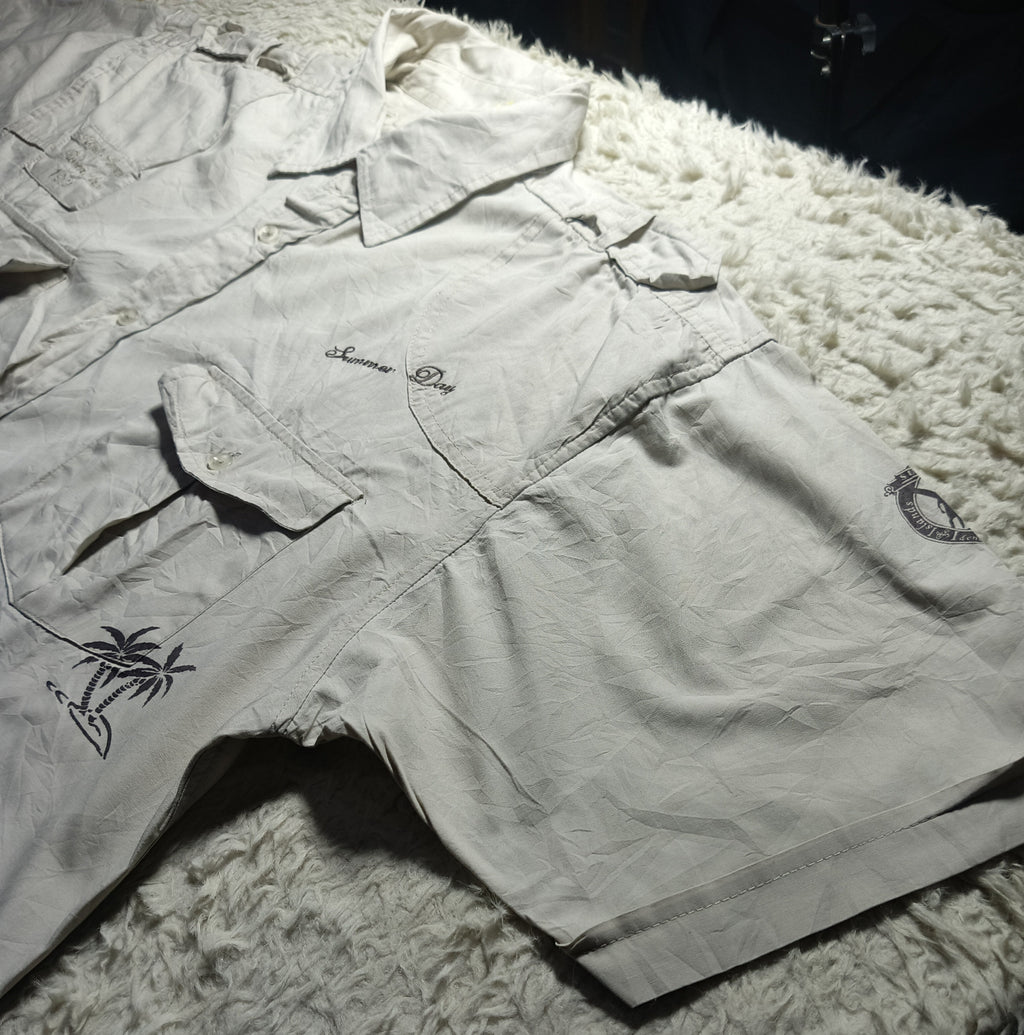 A light beige or off-white short-sleeve shirt and matching shorts laid flat on a white shag rug. The shirt has a small embroidered palm tree design on the lower left side and a small script logo on the chest, while the shorts have a small circular logo on the upper right leg.