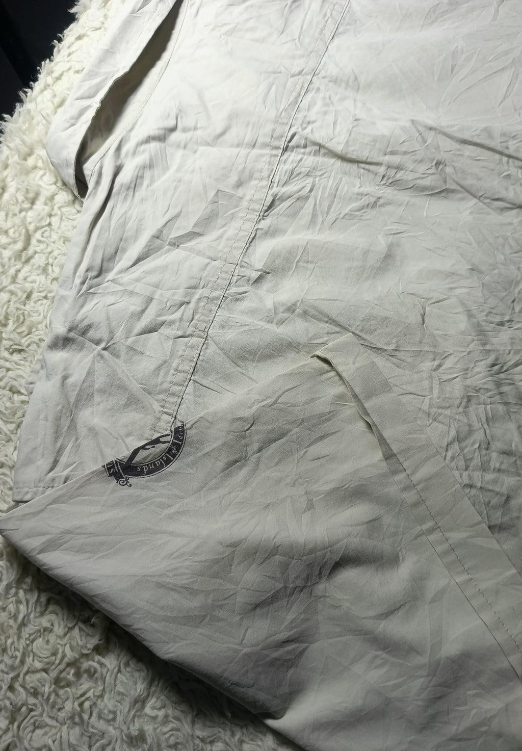 A close-up of a light beige or off-white wrinkled fabric item laid on a white textured rug, with a small dark embroidered logo near the lower left area.