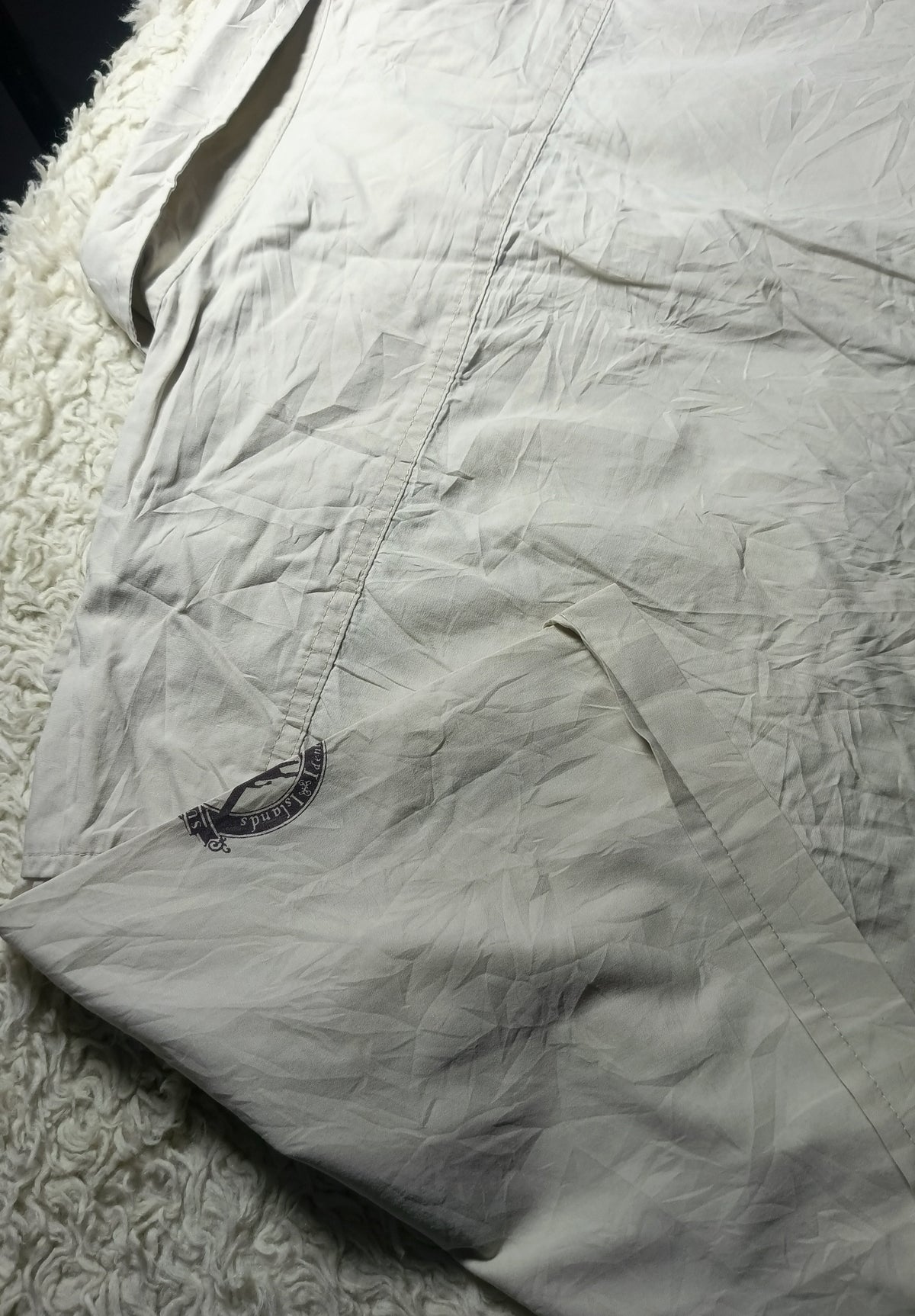 A close-up of a light beige or off-white wrinkled fabric item laid on a white textured rug, with a small dark embroidered logo near the lower left area.
