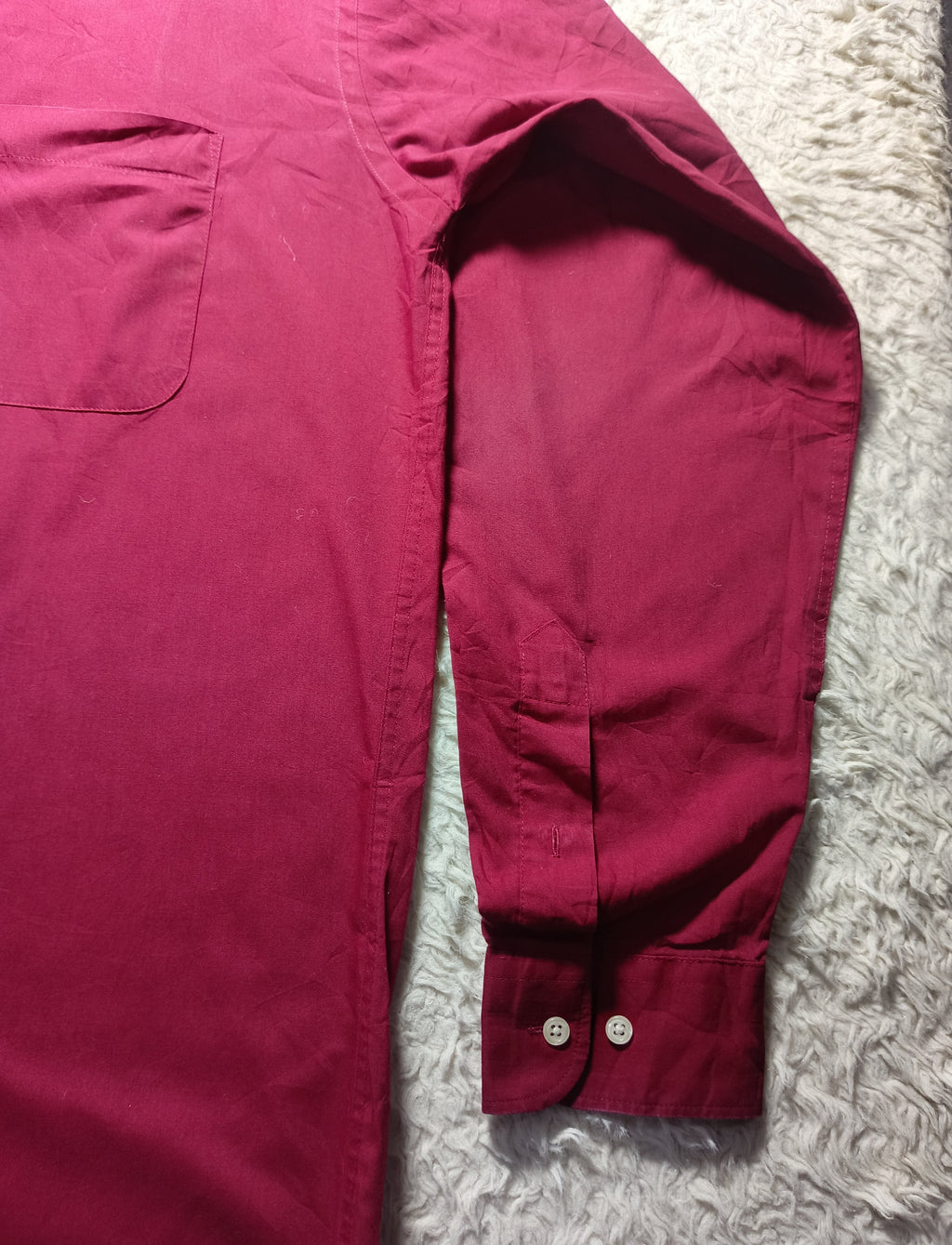A close-up of a long-sleeve button-up shirt laid flat on a textured white rug, showing the chest pocket, sleeve, and buttoned cuff.