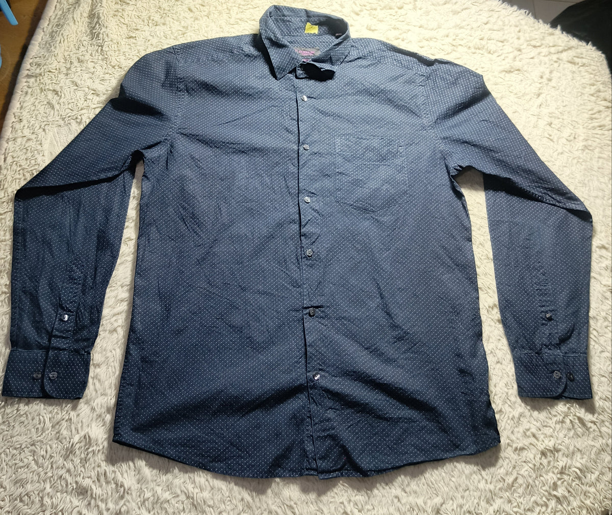 A dark blue long-sleeve button-up shirt laid flat on a white shag rug, with a light box and other items visible in the background.