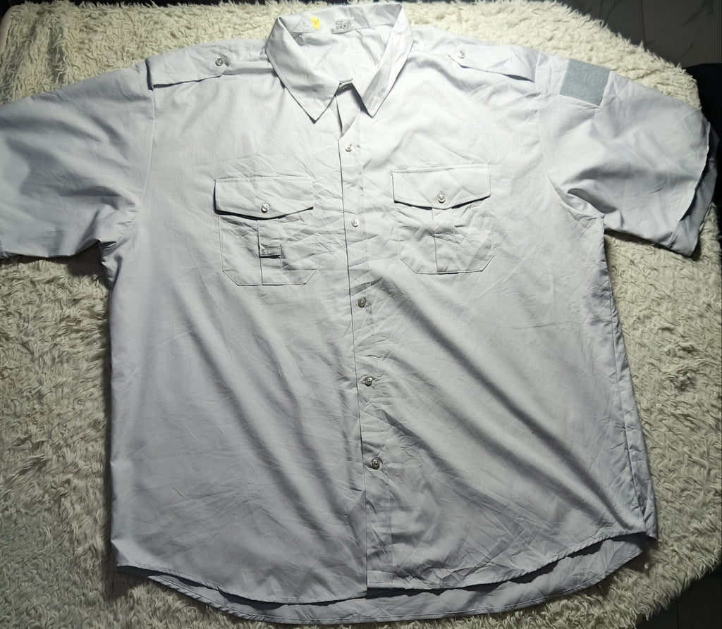 A light gray short-sleeve button-up shirt with two chest pockets, laid flat on a white shaggy blanket.