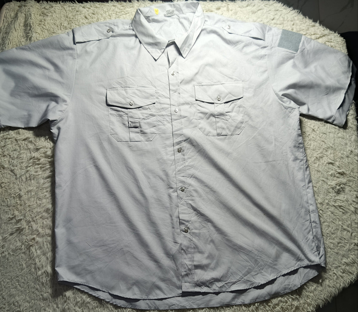 A light gray short-sleeve button-up shirt with two chest pockets, laid flat on a white shaggy blanket.
