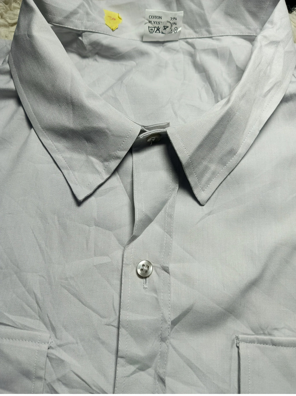 A light gray or silver-gray long-sleeve button-down shirt, shown in a close-up view with the collar and front button visible. The fabric has a slightly crinkled texture, and there is a visible inner neck label and a camera watermark at the bottom.