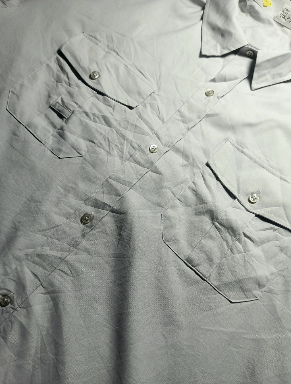 A close-up of a light gray or off-white button-up shirt with multiple chest pockets and visible creases, photographed flat against a dark background. A small brand tag is visible near the upper left chest pocket.