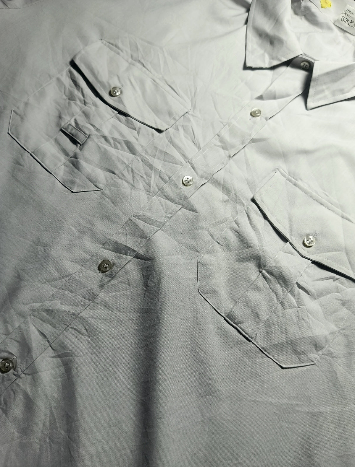 A close-up of a light gray or off-white button-up shirt with multiple chest pockets and visible creases, photographed flat against a dark background. A small brand tag is visible near the upper left chest pocket.
