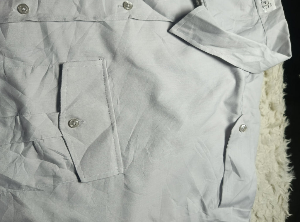 A close-up of a light gray or off-white button-up shirt with a chest pocket and silver-toned buttons, laid flat with visible wrinkles. A white fuzzy textured surface is partially visible on the right side.