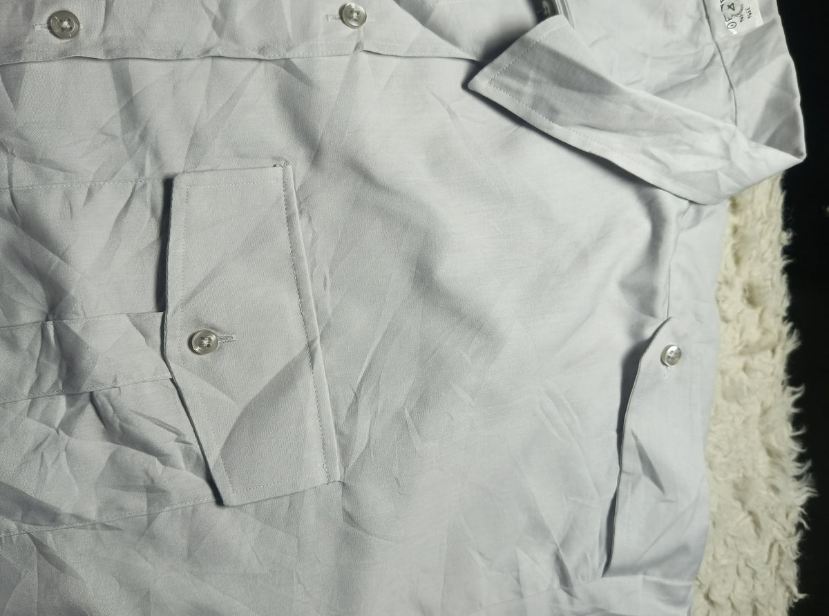 A close-up of a light gray or off-white button-up shirt with a chest pocket and silver-toned buttons, laid flat with visible wrinkles. A white fuzzy textured surface is partially visible on the right side.