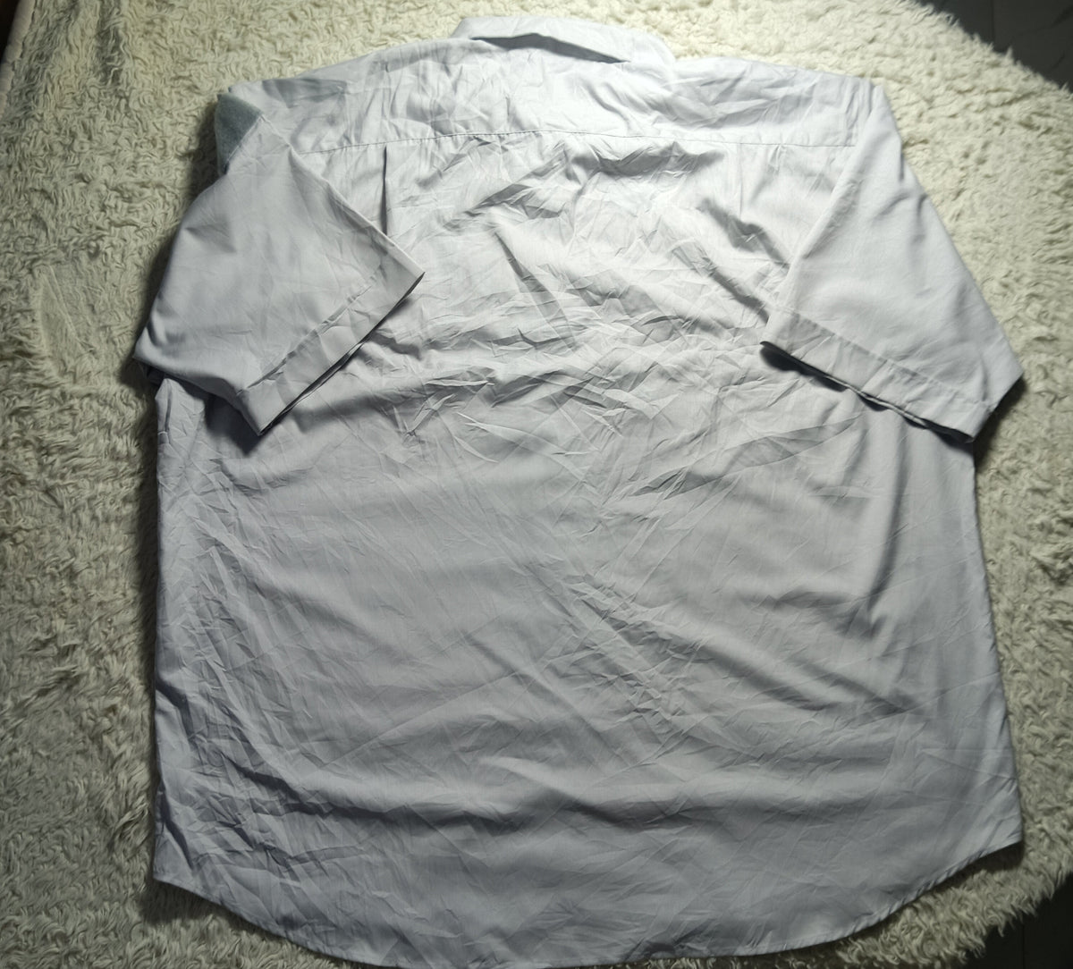 A light gray long-sleeve button-up shirt laid flat on a white shag rug, photographed from above. The shirt is wrinkled and the photo has a casual, amateur feel with a camera watermark at the bottom.