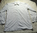 A long-sleeve button-up shirt with thin vertical stripes in light gray and white, laid flat on a shaggy cream rug. The photo is taken in a dimly lit room with wooden chairs and a light stand visible in the background.