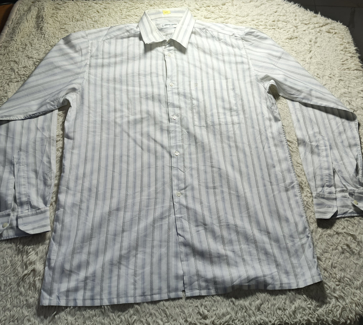 A long-sleeve button-up shirt with thin vertical stripes in light gray and white, laid flat on a shaggy cream rug. The photo is taken in a dimly lit room with wooden chairs and a light stand visible in the background.
