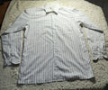 A long-sleeve button-up shirt with thin vertical stripes in light gray and white, laid flat on a shaggy cream rug. The photo is taken in a dimly lit room with wooden chairs and a light stand visible in the background.